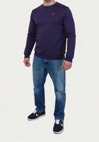 Fialová mikina s dlouhými rukávy a kulatým výstřihem, zdobená malým červeným emblémem. Nošena s světle modrými džíny a tmavými teniskami.