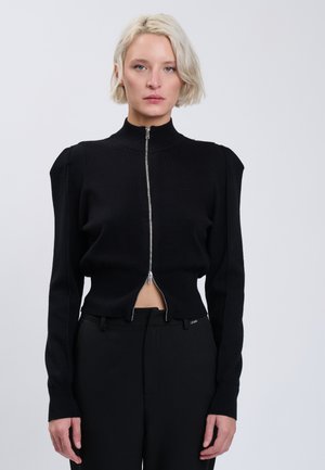 Blonde Frau trägt einen schwarzen Reißverschluss-Pullover mit aufgepufften Schultern und schwarze Hose, steht vor einem einfarbigen hellen Hintergrund.