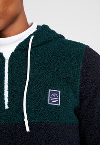 Hoodie en polaire vert foncé et bleu marine. Comprend un zip demi-long, un tissu texturé, un cordon de serrage réglable et un patch logo sur la poitrine.