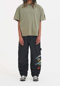 Olivgrön bomullströja med korta ärmar, kombinerad med svarta cargobyxor som har färgglad drakbroderi och en avslappnad passform.