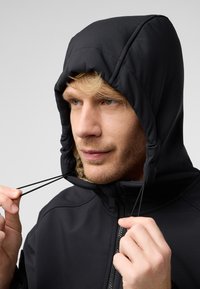 Veste à capuche noire avec un intérieur texturé. Elle dispose de cordons de serrage ajustables et d'un zip frontal, mettant en avant un design moderne et une fonctionnalité.
