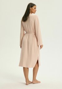 Jasnoróżowy szlafrok z długimi rękawami, związywany w pasie, o długości do połowy łydki i z bocznymi rozcięciami, wykonany z miękkiej, gładkiej tkaniny.