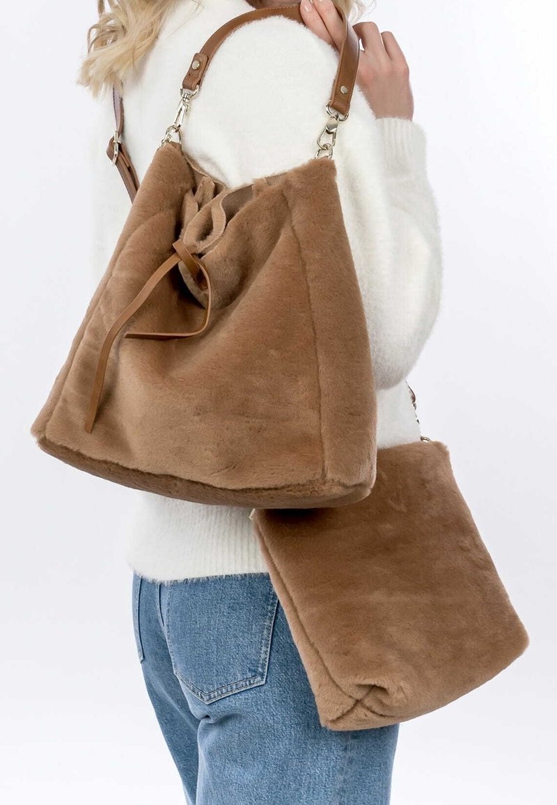 Personne portant un pull clair et un jean, portant deux sacs en fausse fourrure marron avec des sangles en cuir à l'épaule et au bras.