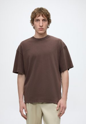 Jeune homme aux cheveux bouclés portant un t-shirt marron ample et un pantalon beige clair, debout devant un fond blanc uni.