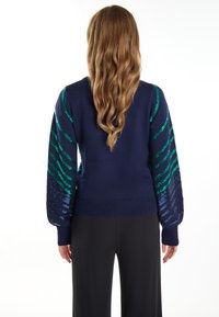 Marineblauer Pullover mit grünen und blauen Paillettenstreifen an den Ärmeln; gerippte Bündchen und Saum; strukturiertes Strickmaterial. Model trägt eine dunkle Hose.