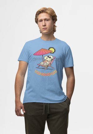 Ung mand iført lyseblå T-shirt med Snoopy, der læner sig tilbage under en lyserød paraply og solgrafik, med påskriften "Det gode liv."