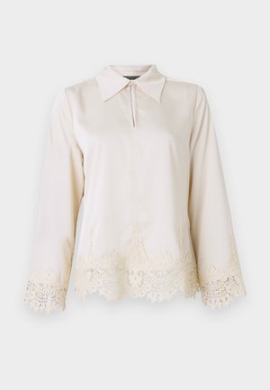 Blouse en crème avec un col, fermeture éclair à l'avant, et manches longues ornées d'une délicate dentelle au bas et aux bords des manches.