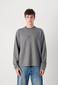 Pilkas megztinis su apvaliu iškirpimu, su iškilniuoju „CK“ logotipu ant krūtinės. Derinamas su šviesiai mėlynomis džinsinėmis kelnėmis.