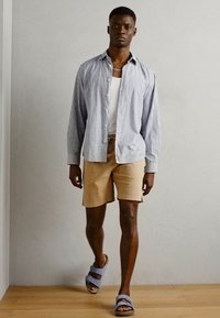 Lyseblå og hvid stribet langærmet skjorte, hvid tanktop, brune shorts og blå slidesandaler. Enkel, afslappet sommeroutfit.