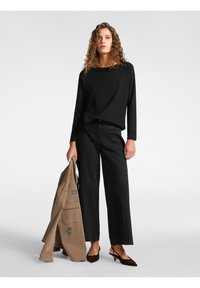 Top nero a maniche lunghe, vestibilità rilassata, abbinato a pantaloni neri a gamba larga. Il modello tiene in mano una giacca beige con ricami e indossa tacchi marroni scuri.