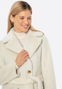 Abrigo de color claro y peludo con cuello con muesca, cierre de botón y correa atada. Presenta una textura suave y un diseño de ajuste holgado.