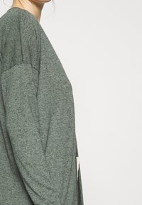 Długi kardigan na długim rękawie w odcieniu zieleni melanżowej, o luźnym kroju, z delikatną fakturą i bocznym rozcięciem zapewniającym swobodę ruchów. Brak widocznych elementów metalowych.