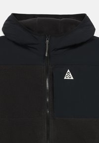 Jachetă fleece neagră cu glugă, fermoar complet pe față și buzunar pe piept cu fermoar, având un logo triunghiular alb cu literele "acg".