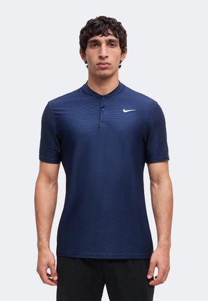 Homme debout face à la caméra, portant un polo Nike bleu marine à manches courtes avec deux boutons et un pantalon noir.