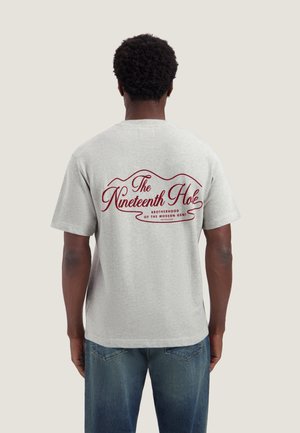 Man draagt een lichtgrijs T-shirt met rode tekst "The Nineteenth Hole Brotherhood of the Modern Gent" op de rug en blauwe spijkerbroek.