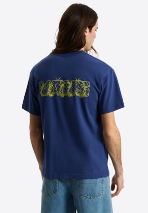 Man met lang bruin haar draagt een blauw Vans T-shirt met gele grafische tekst op de rug en lichtblauwe jeans, met de rug naar de camera.