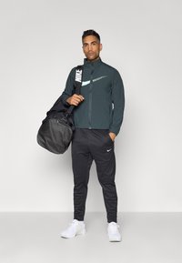 Tummanvihreä vetoketjullinen takki, jossa on vaaleansininen Nike-logo, mustat verkkohousut, joissa on valkoinen Nike-logo, valkoiset tennarit ja musta viikonloppulaukku.