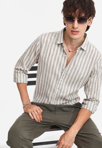 Camicia di lino a righe beige e verde, senza colletto e con bottoni, maniche arrotolate. Abbinata a pantaloni verde oliva e occhiali da sole grigi.
