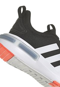 Černé síťované sneakers s bílými pruhy, silnou bílou podrážkou s průsvitným modrým polstrováním a červeným gumovým vzorkem na patě.