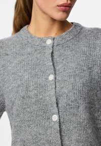 Pilkas megztas kardiganas su apykakle, turintis tekstūruotą raštą ir keturis baltus mygtukus priekyje, skirti užsegti.