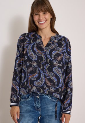 Femme souriante portant un chemisier à manches longues à motif cachemire bleu et marron avec des rayures aux poignets et un jean en denim bleu.