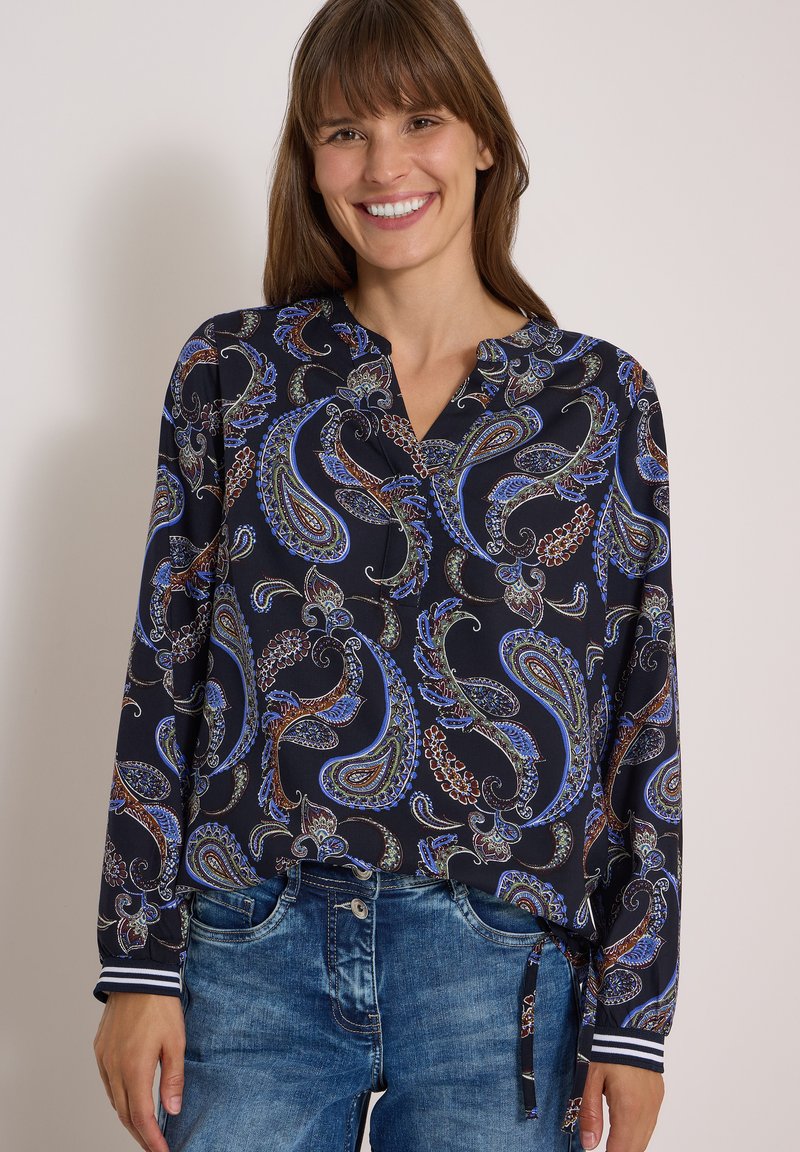 Femme souriante portant un chemisier à manches longues à motif cachemire bleu et marron avec des rayures aux poignets et un jean en denim bleu.