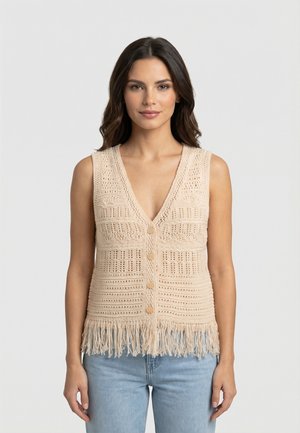 Donna con capelli lunghi e scuri che indossa un gilet senza maniche beige lavorato a maglia con bottoni e frange, abbinato a jeans azzurri, in piedi davanti a uno sfondo bianco.