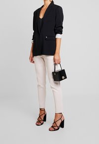 Blazer noir à manches longues, pantalon ajusté de couleur claire, talons hauts noirs à lanières et un petit sac à main noir avec une finition texturée.