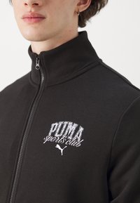 Giacca nera con zip, caratterizzata da un alto colletto, una texture morbida e un logo ricamato bianco "PUMA sports club" sul lato sinistro del petto.