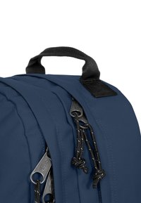 Mochila azul marino con una tela texturizada, que cuenta con un asa superior y tiradores de cremallera negros con cordones trenzados para un acceso fácil.