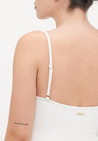 Débardeur blanc à fines bretelles avec un motif floral texturé. Présente des éléments en or sur la bretelle et un petit logo doré.
