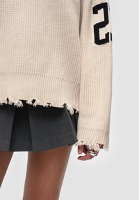 Beige gestrickter Pullover mit ribbeligem Halsausschnitt und fransigem Saum. Schwarze Nummer "2" auf dem Ärmel. Das Model trägt einen grauen plissierten Rock.
