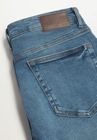 Jeans de mezclilla azul con un parche de cuero marrón en la cintura. Presenta un diseño clásico de cinco bolsillos y detalles de costura en los bolsillos.