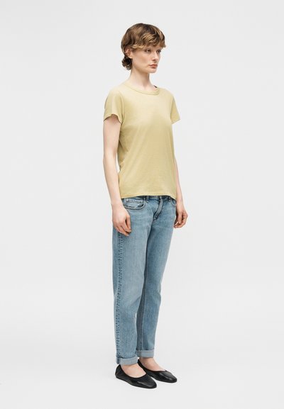 Νέος/νέα με κοντά μαλλιά, φοράει μπεζ μπλούζα, ανοιχτό μπλε τζιν με γυρισμένα μπατζάκια και μαύρα φλατ παπούτσια, στέκεται μπροστά σε απλό φόντο.