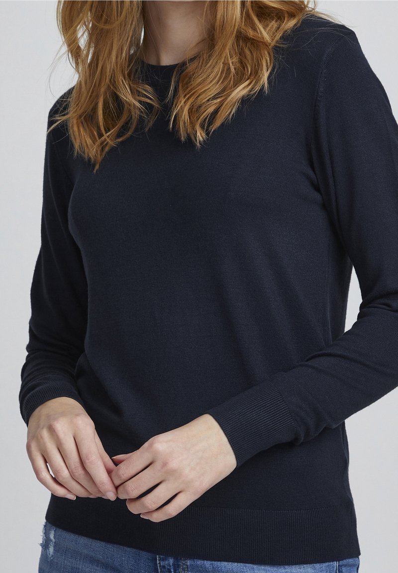 Femme portant un pull en maille à manches longues bleu marine foncé avec poignets et ourlet côtelés, associé à un jean bleu déchiré.