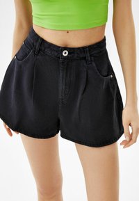 Shorts en denim noir à taille haute, avec des poches avant, une fermeture à bouton et une texture lisse. Complétés par un haut ajusté vert.