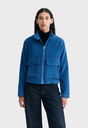 Femme portant une veste zippée en velours côtelé bleu avec des poches à rabat boutonnées et un pantalon noir, debout devant un fond clair uni.