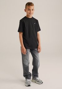 Svart kortärmad t-shirt med rund nacke och en liten vit logotyp, i kombination med grå raka jeans och grå sneakers med vita detaljer.