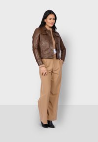 Veste cropped en cuir marron avec fermeture à boutons, deux poches poitrine et un col, associée à un pantalon beige taille haute et des bottes noires.
