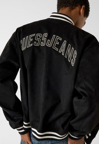 Bomber nero con texture liscia, caratterizzato da "GUESS JEANS" ricamato sulla schiena e da polsini e collo a costine a righe.