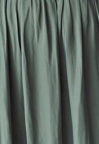 Zelená plisovaná látka s hladkým povrchem. Materiál vykazuje jemný lesk a měkce se splývá, vytváří jemné záhyby.