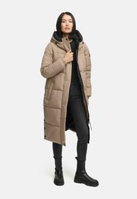 Lange beige puffer jas met capuchon, voorzien van quilted panelen, voorrits en drukknoopsluitingen. Gedirigeerd over een zwarte top en zwarte jeans.