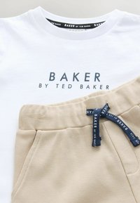 Witte katoenen T-shirt met "BAKER BY TED BAKER" in donkerblauwe tekst, gecombineerd met beige joggingsbroek met een marineblauwe trekkoord.
