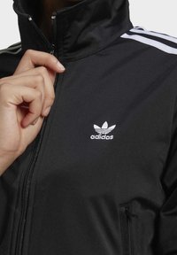 Svart zip-up jacka av mjukt tyg, med hög krage, vita ränder på axlarna och en liten vit logotyp på bröstet.