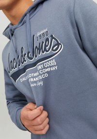 Szary hoodie z wyraźnym białym i granatowym logo, które łączy tekst pisany kursywą i blokowy, charakteryzujący się miękką fakturą i regulowanym kapturem ze sznurkiem.