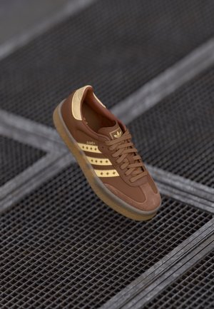 Brązowy sneaker Adidas Samba z beżowymi paskami i wzorami gwiazdek, unoszący się nad metalową kratownicą podłogi.