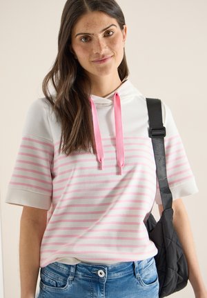 Jeune femme aux longs cheveux bruns portant un sweat à capuche à manches courtes blanc à rayures roses et un sac à bandoulière noir.