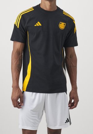 Homme portant un maillot de football jamaïcain noir et jaune avec le logo Adidas et un short blanc avec des bandes noires Adidas.