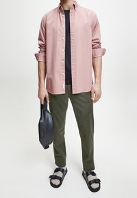 Camicia button-up rosa chiaro sopra una maglietta nera, pantaloni verde oliva, sandali neri e una borsa nera con logo. Outfit casual.