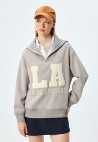 Jasnoszary sweatshirt z dużym białym napisem "LA", prążkowanym kołnierzem i mankietami, z zamkiem błyskawicznym przy szyi, noszony z białą koszulą i granatową spódnicą.